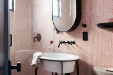 a small blush bathroom clad with pink penny tiles, a shower space, a creative sink on a stand, an oval mirror and black fixtures