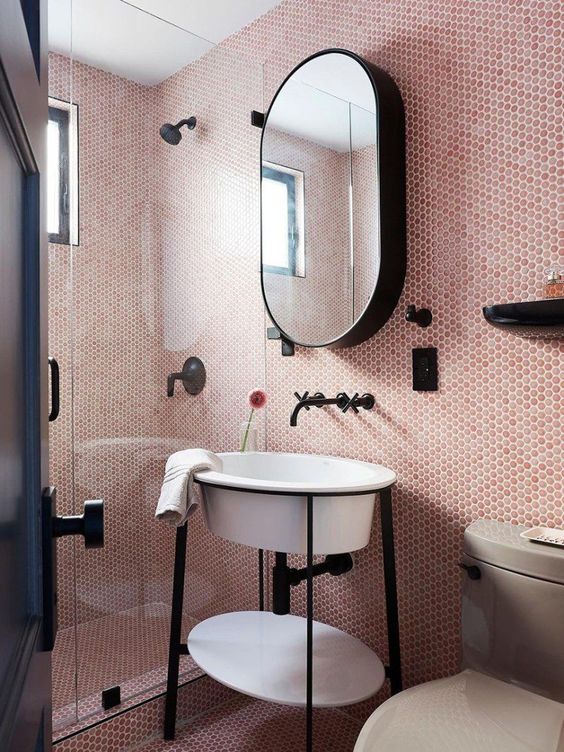 a small blush bathroom clad with pink penny tiles, a shower space, a creative sink on a stand, an oval mirror and black fixtures