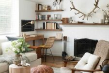 a stylish modern living room done in neutrals, with a vintage fireplace, chic and comfy furniture, a small workspace in the corner, floating shelves and antlers