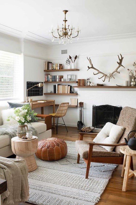 a stylish modern living room done in neutrals, with a vintage fireplace, chic and comfy furniture, a small workspace in the corner, floating shelves and antlers