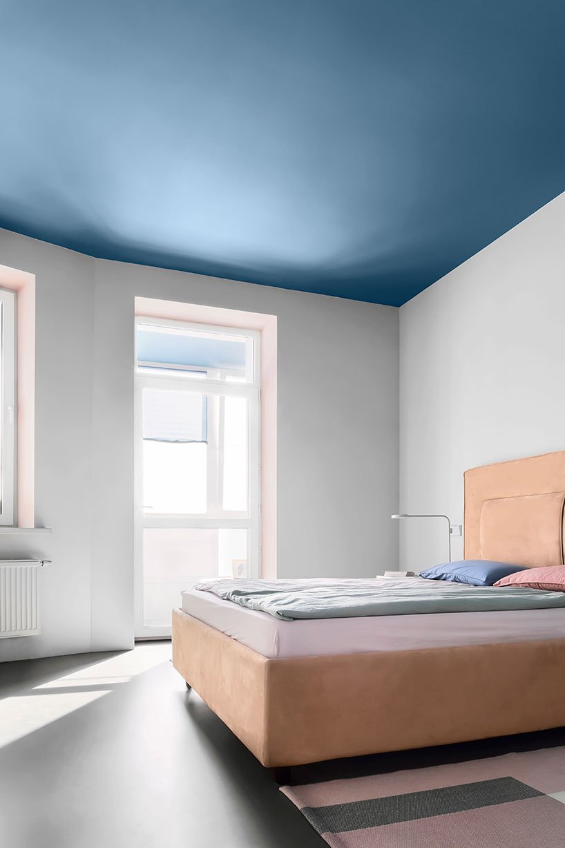 The bedroom is done with a blue ceiling, a peachy pink upholstered bed, sconces and a large window to fill it wiht light