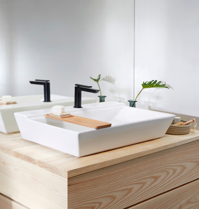 The washbasin has a geometric shape, a black matte tap and a little wooden tray