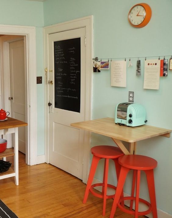 A tiny colorful breakfast bar with a wall mounted table, red chairs and a blue toaster