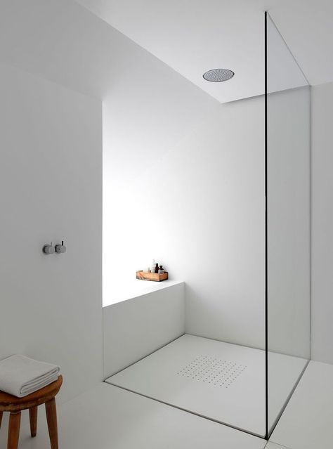 An ultra minimalist bathroom in white with a glass shower and a wooden stool