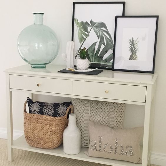 a summer console with artworks with a pineapple and tropical leaves, a potted plant, a basket with a pineapple pillow and a tropical feel