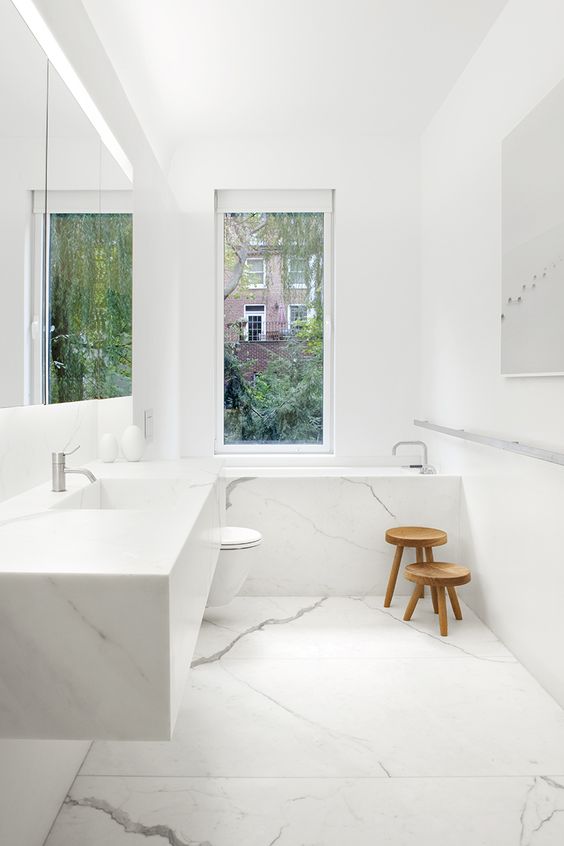 a minimalist white marble bathroom with a narrow window and a couple of wooden stools