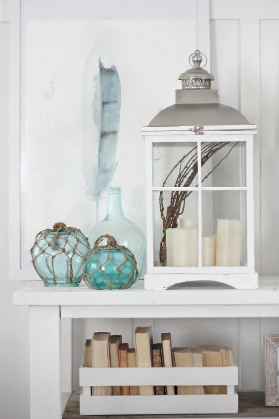 a beachy console with an oversized lantern with candles, floats, a feather artwork and a crate with books