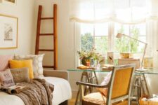 a cozy guest bedroom with a sofa with pillows, a glass trestle desk, an orange chair, a printed rug and a ladder as decor