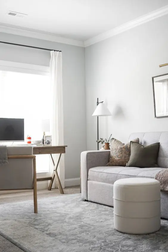 a small neutral guest room with a grey sofa, a trestle desk, a grey chair, a pouf and a floor lamp plus some decor