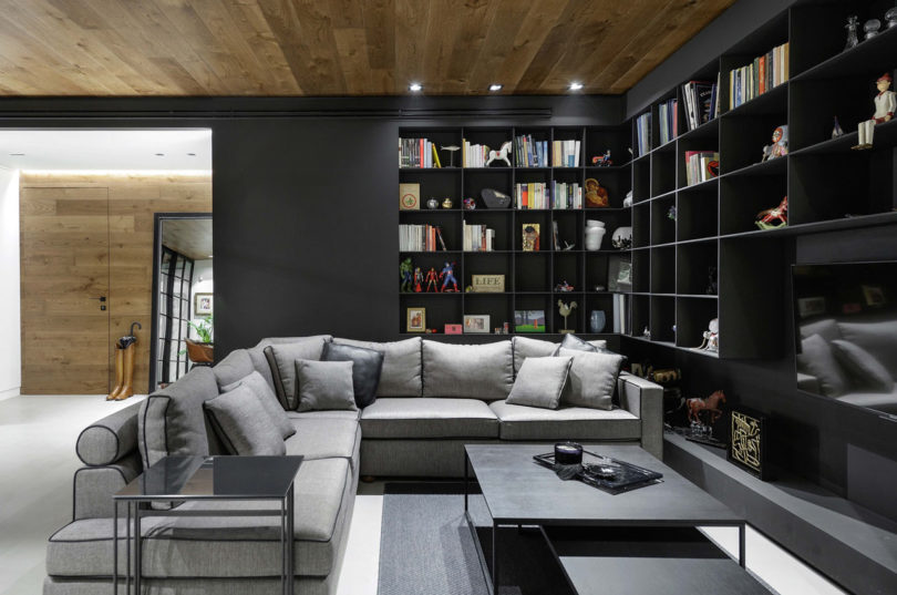 The living room features a lot of black bookshelves, a large corner sofa and some coffee tables plus a wood clad ceiling