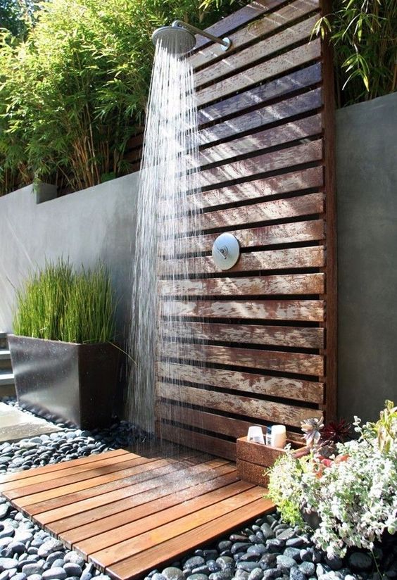 a spa outdoor shower of wooden planks with pebbles on the ground and greenery around