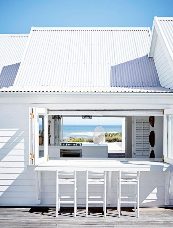 An all white beach house with a folding window, a white bar counter and stools and the same window on another wall to see the views