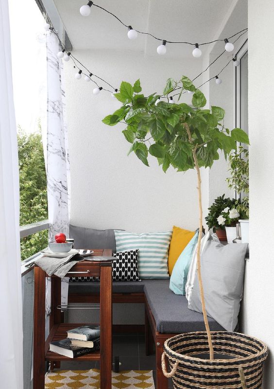 A small cozy balcony with an L shaped upholstered bench, a small coffee table and potted plants