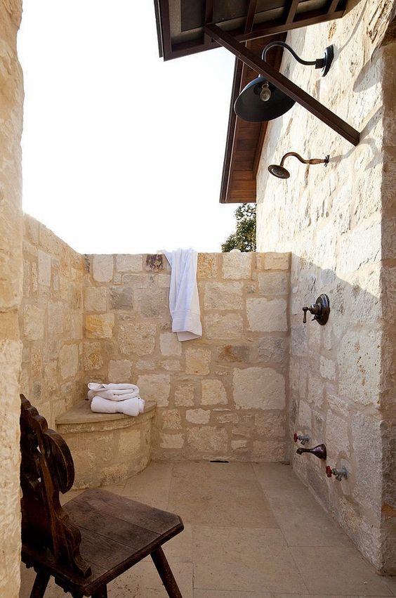 A cozy outdoor rustic shower fully clad with stone, with a little built in storage item and a chair