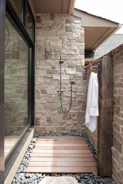 a small yet stylish outdoor shower clad with stone and bricks, a wooden deck and pebbles on the ground