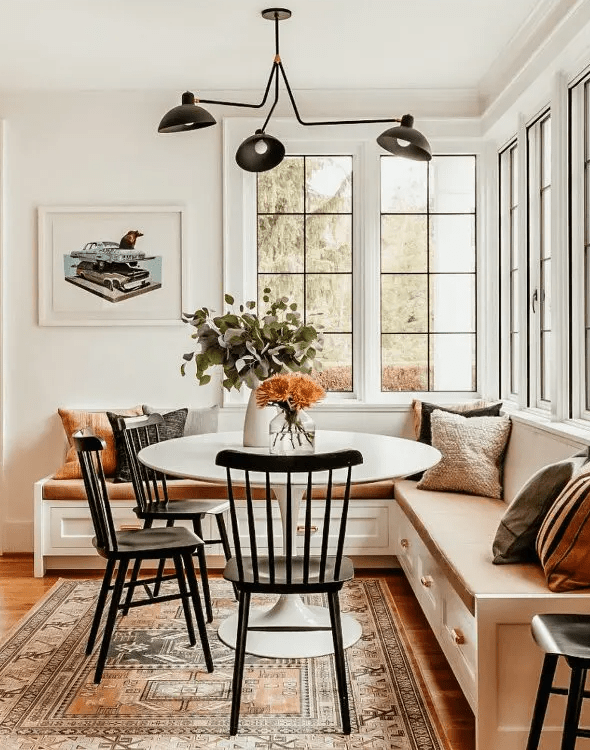 a cozy modern farmhouse breakfast space with a lovely corner storage seating, a round table, black chairs, a black chandelier and some pillows