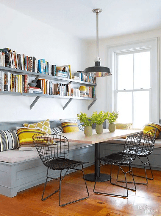 a farmhouse dining space with a grey banquette with colorful pillows, a table on a metal leg, open shelving, black metal chairs