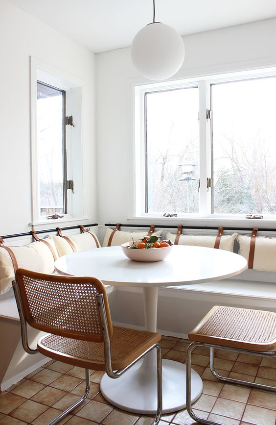 a modern dining space with a banquette seating, a white table, cane chairs, a pendant lamp and some cushions