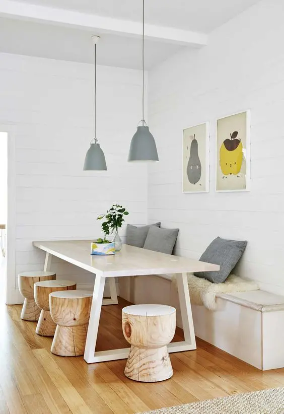 a neutral dining room with a banquette seating, a table, wooden stools, pendant lamps and some artwork