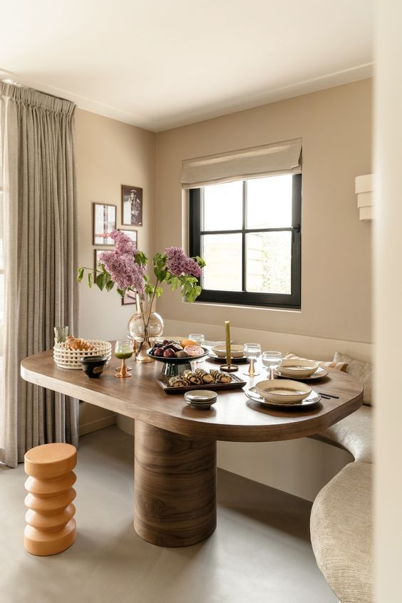 a neutral modern dining room with a banquette seating, a stained table, chic gallery wall, a stool and neutral drapes