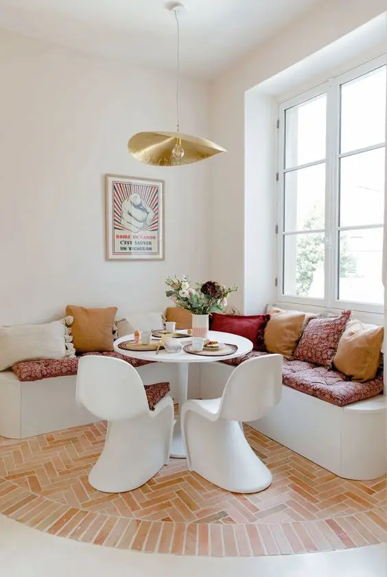 a pretty dining nookw ith a corner banquette seating, a small table and white chairs, a gold pendant lamp and lots of pillows