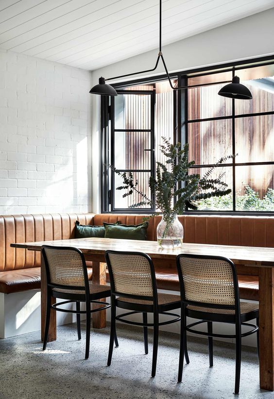 a refined and elegant modern dining room with a large leather banquette seating, a stained table and cane chairs, a black chandelier and greenery
