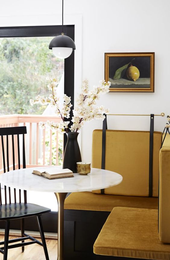 a refined vintage dining space with a mustard banquette seating, a round table, a black chair, a pendant lamp and a vintage artwork
