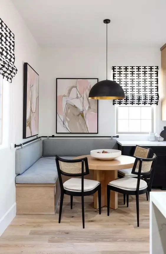 a stylish modern dining nook with a banquette seating, a stained table, cane chairs, a black pendant lamp and oversized artwork
