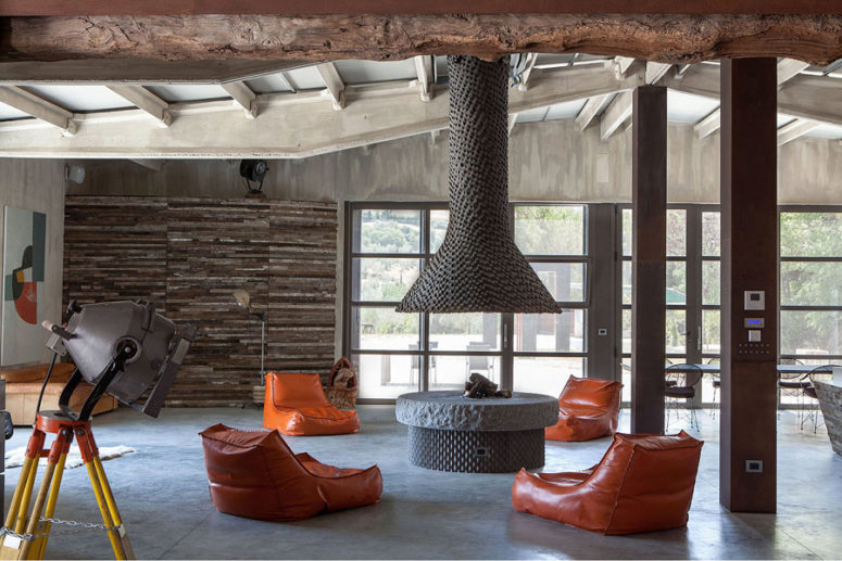 The main layout includes a living and dining space plus a kitchen, it's done with concrete, weathered wood and metal, a large hearth with orange chairs is a centerpiece