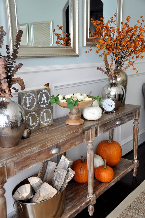 a fall console with orange and white pumpkins, fall leaves and dried blooms and firewood in a metal bathtub