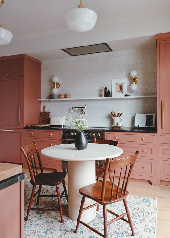 a pink kitchen with a black counterop, a white square tile backsplash, an open shelf with decor and lovely sconces