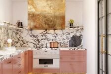 a refined and glam kitchen with pink cabinets, a marble backsplash and countertops, a gold hood looks wow