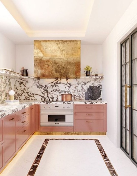 A refined and glam kitchen with pink cabinets, a marble backsplash and countertops, a gold hood looks wow