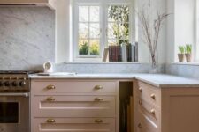 an elegant and chic kitchen done in blush, with white stone countertops and brass handles, a white stone backsplash and some potted plants