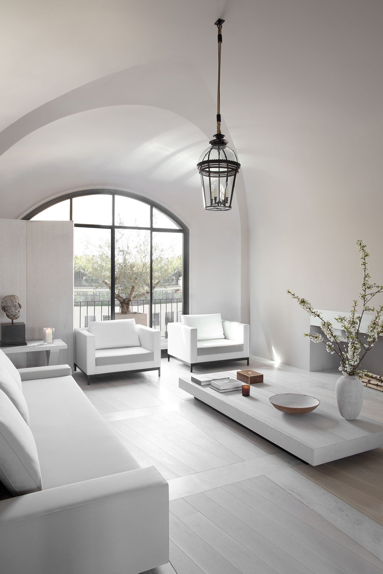 The living room is a light filled space with white furniture and much light coming through an arched window