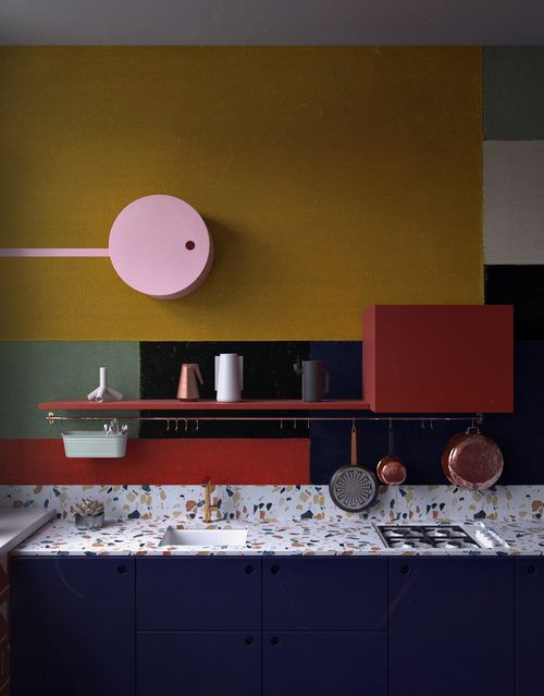 a dark blue kitchen with a terrazzo countertopp, brass fixtures and postal red shelf plus green and yellow color blocking