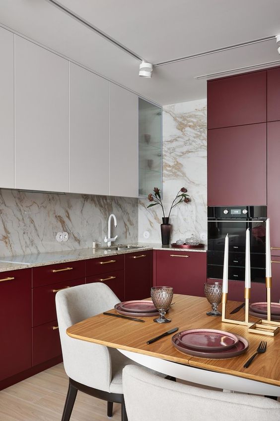 A beautiful burgundy and white eat in kitchen with white stone countertops, a table and neutral chairs and built in appliances