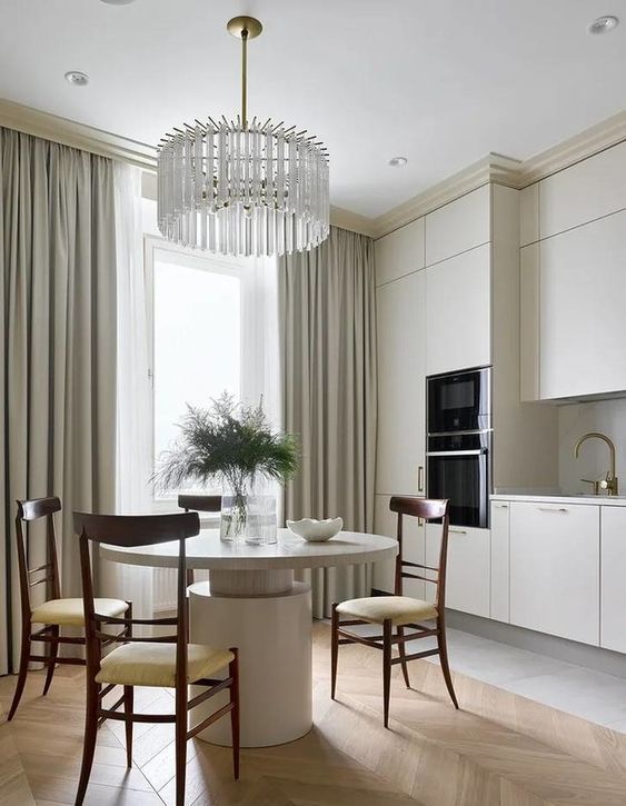A sophisticated neutral eat in kitchen with creamy cabinets, a round table, rich stained chairs and a crystal chandelier