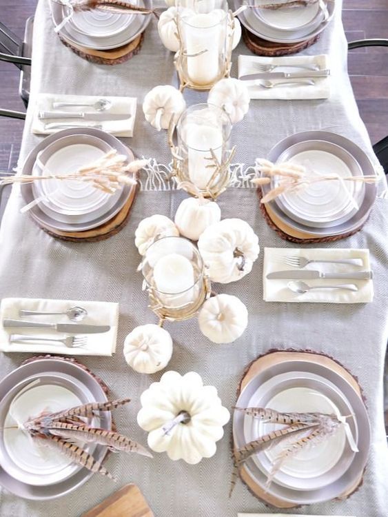 a chic neutral table setting with a grey tablecloth, white pumpkins and candles and feathers for each place setting