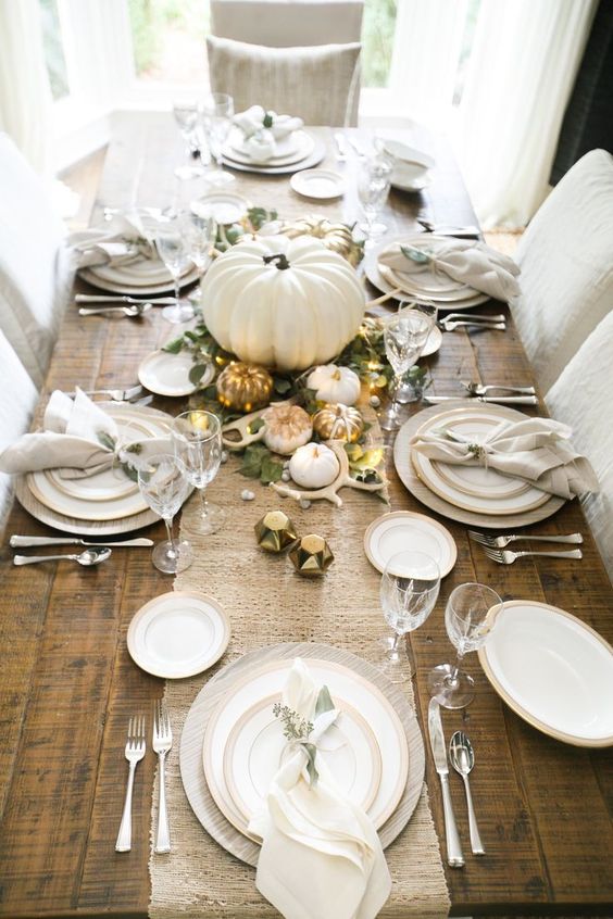 a table runner of white burlap, little white and gilded pumpkins, greenery, antlers and LEDs on an uncovered table