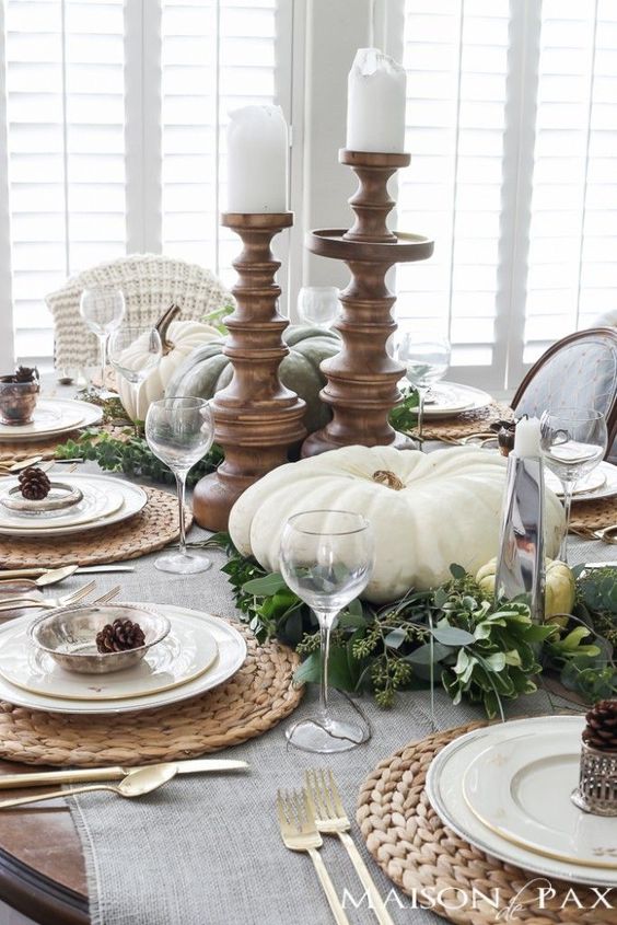 a neutral table setting with large white pumpkins and candles, pinecones, fresh greenery and woven placemats