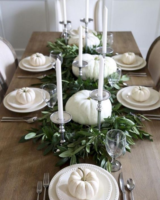 a neutral tablescape with white pumpkins, plates and candles and a fresh greenery table runner in the center