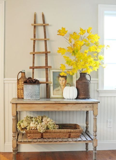 a rustic console with a fall leaf in a metal vase, pinecones, a fake pumpkin, hydrangeas in a wicker tray and artworks