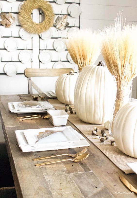 a neutral rustic table setting with large pumpkins, wheat, gilded cutlery and white plates, a wheat wreath on the wall