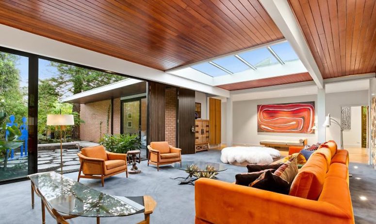The living room features bold orange furniture, catchy glass tables, an entrance to an outdoor space and a large skylight