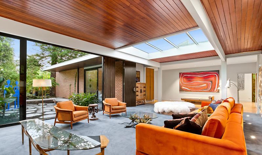 The living room features bold orange furniture, catchy glass tables, an entrance to an outdoor space and a large skylight