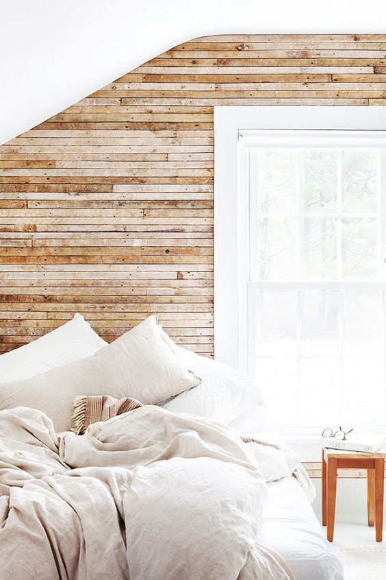 A light filled bedroom with a wood clad wall, neutral linens and a large window