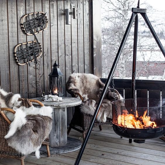 A cabin style terrace with wicker chairs and a wooden table, faux fur and a large suspended fire bowl