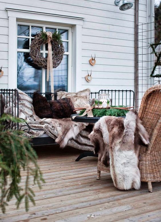a cozy winter terrace with a metal bench, wicker chairs, faux fur throws and pillows, evergreens and candle lanterns
