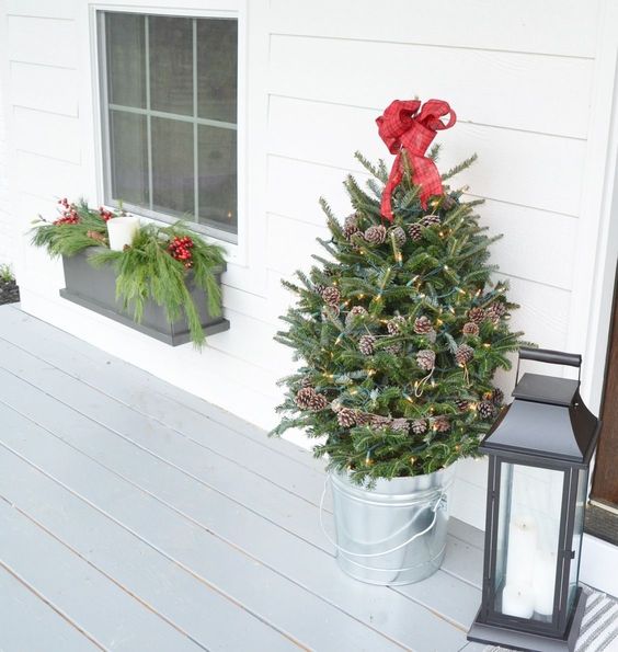 a small holiday tree decorated with lights, pinecones and a red bow on top in a galvanized bucket for a porch